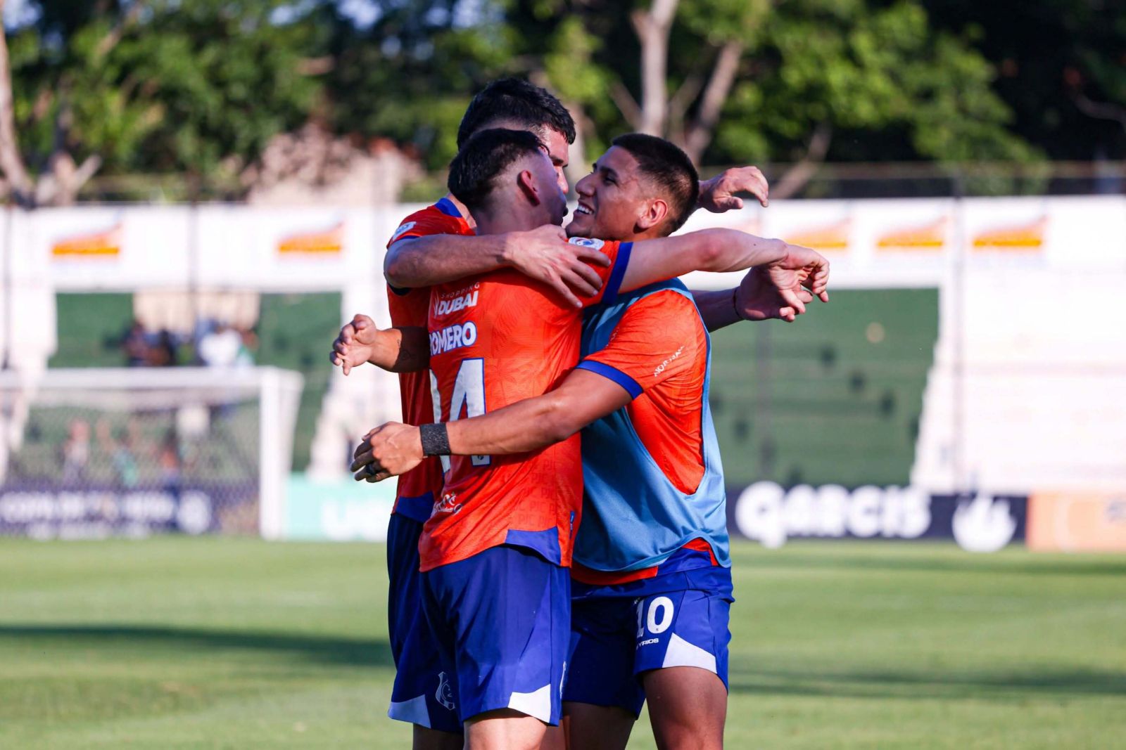 Tres jugadores del 2 de Mayo, con uniforme naranja y short azul, se abrazan en el césped para celebrar