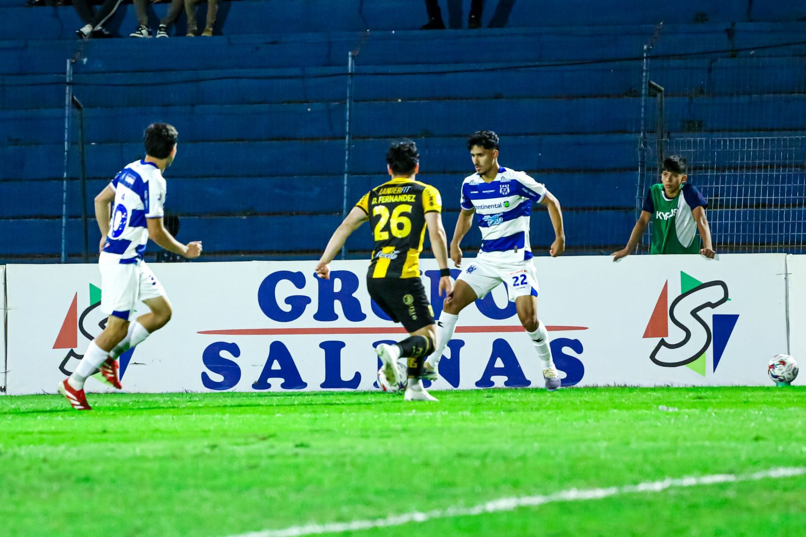 Lance noturno de jogo do 2 de Mayo, com dois jogadores de camisa listrada azul e branca cercando um adversário de listras amarelas e pretas junto à faixa de publicidade à beira do campo