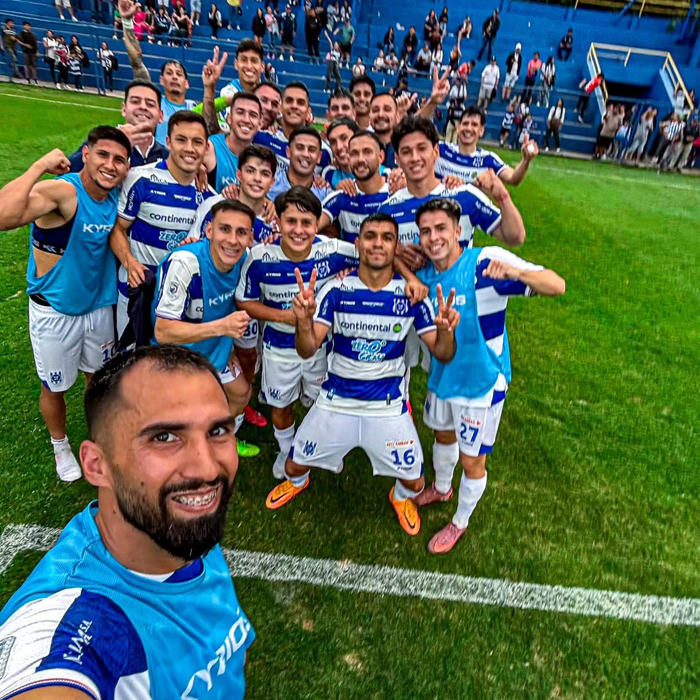 Jugadores del 2 de Mayo reunidos en el césped para una selfie de festejo después del partido, con camiseta a rayas azul y blanca, y la hinchada en la tribuna al fondo.