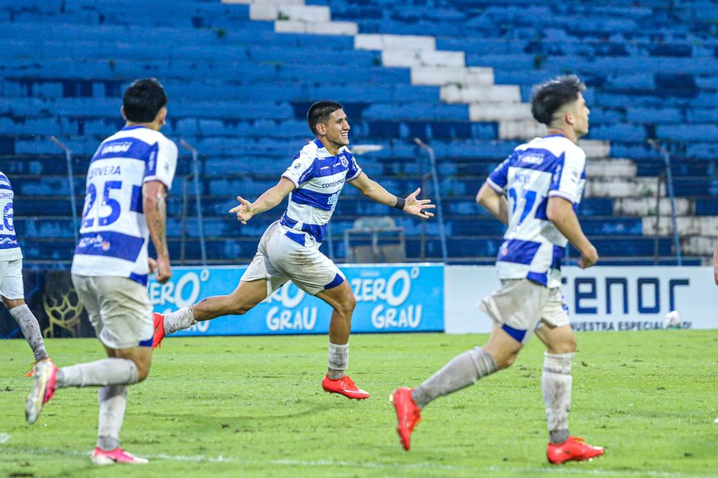 Jogadores do 2 de Mayo, de camisa listrada azul e branca, correm pelo gramado comemorando um gol diante da arquibancada vazia