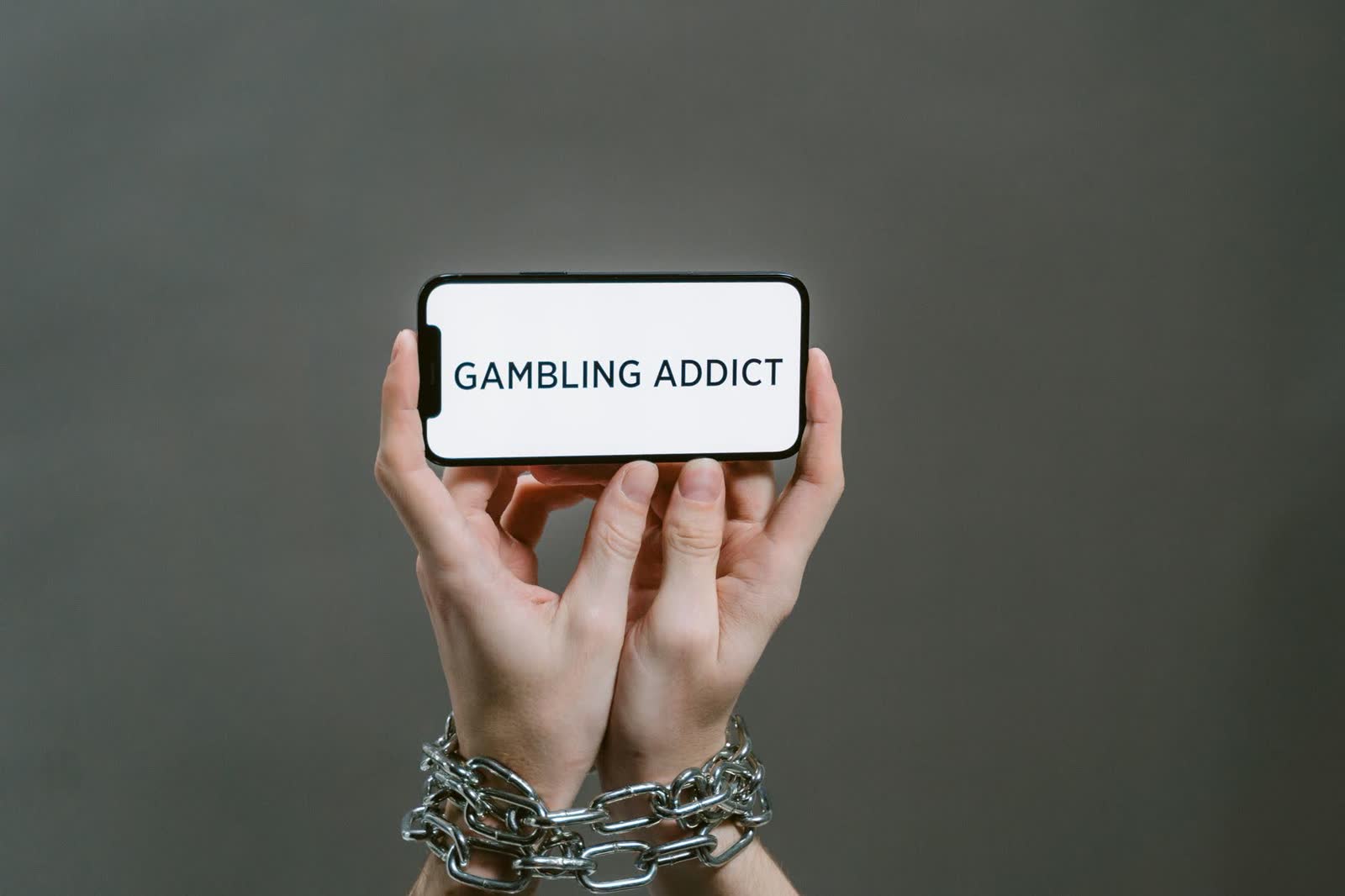 Mãos acorrentadas seguram um celular na horizontal com a frase Gambling Addict na tela, sobre fundo cinza