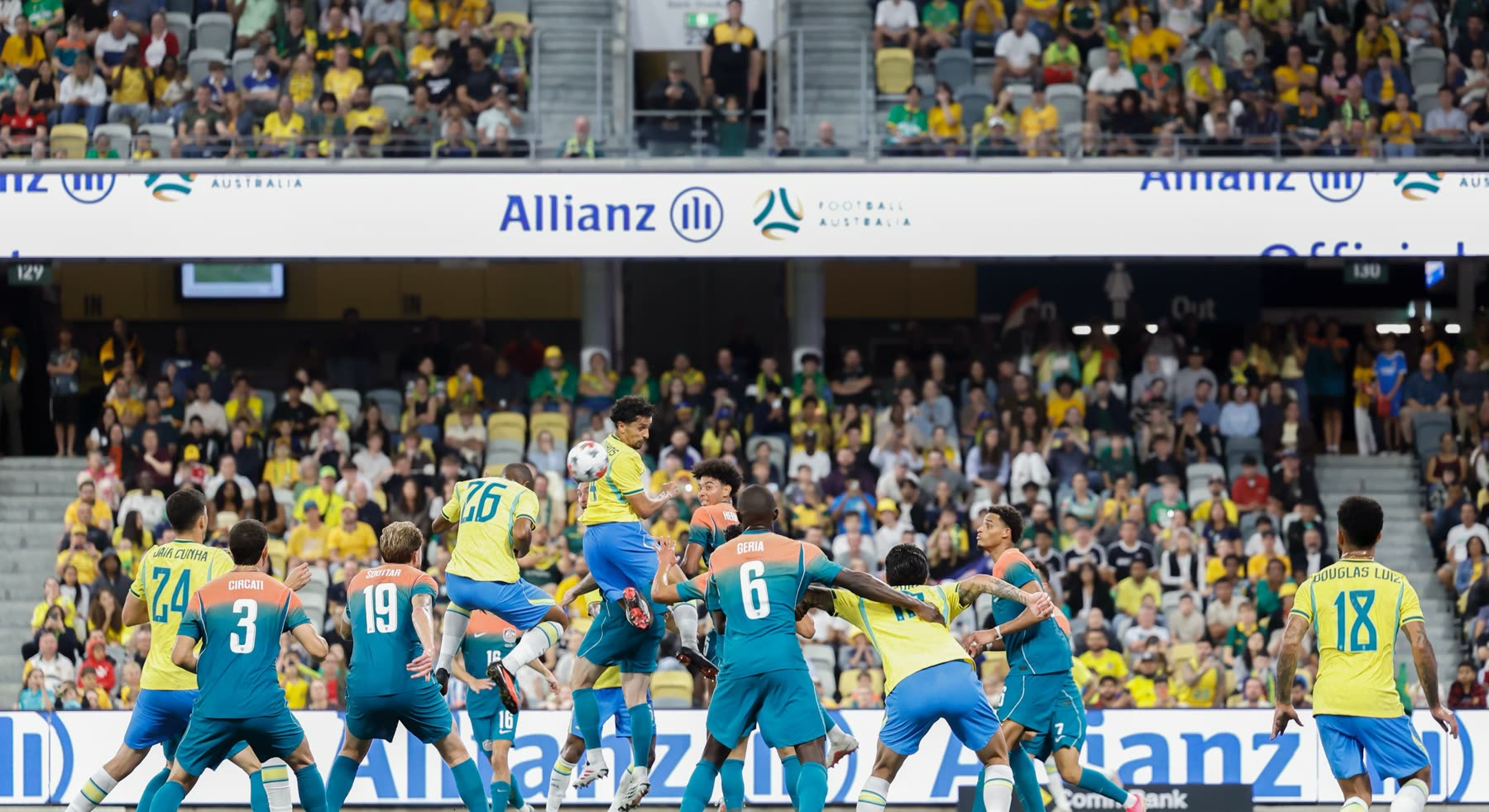 Jugadores de Brasil, de amarillo, y de Australia, de verde agua y naranja, disputan una pelota alta dentro del área, con la tribuna llena al fondo
