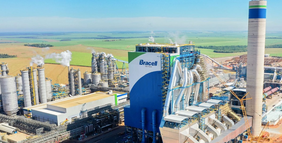 Vista aérea de uma fábrica de celulose da Bracell, com torres, esteiras e a chaminé alta cercadas por plantação