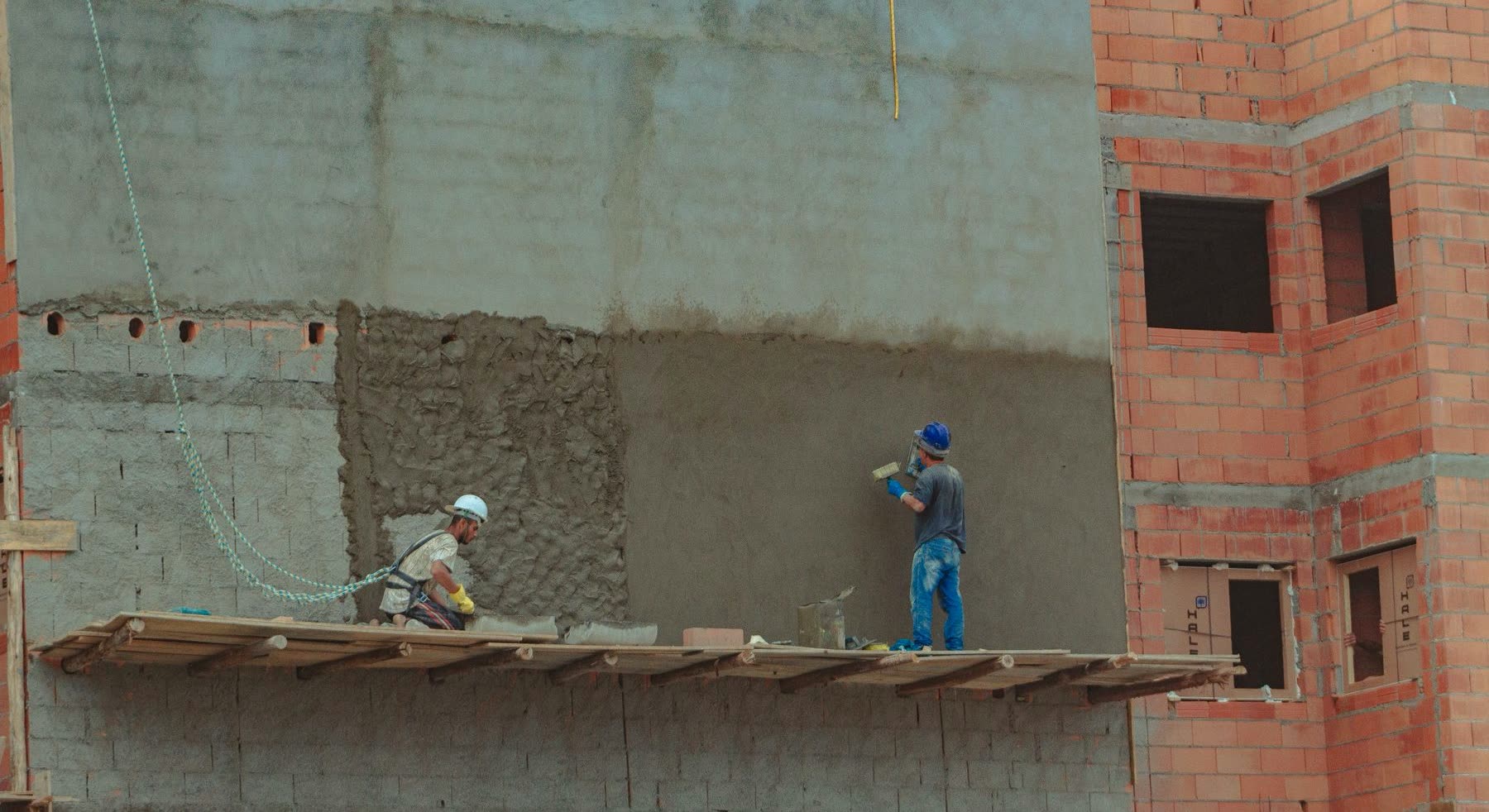 Dos obreros de la construcción revocan la pared de un edificio de ladrillos, sobre un andamio de madera