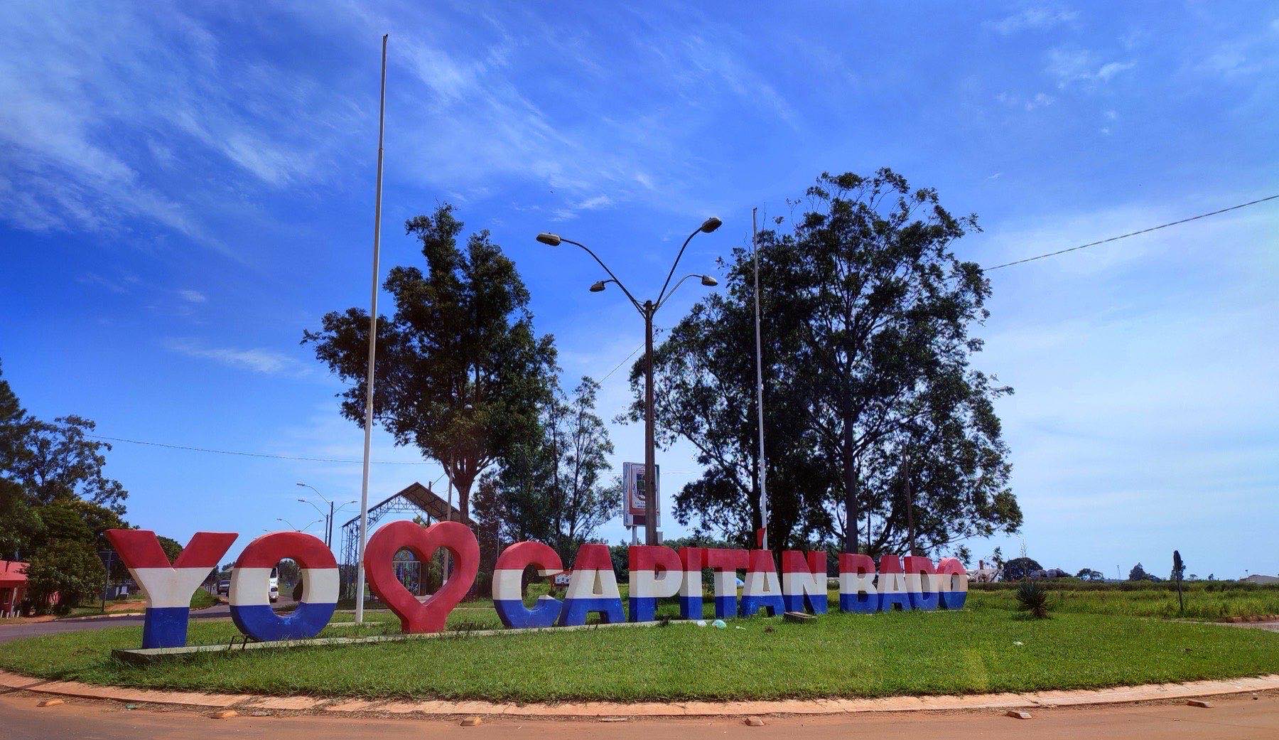 Cartel con las palabras YO AMO CAPITÁN BADO en letras grandes pintadas con los colores de la bandera paraguaya, en un cantero de césped en la entrada de la ciudad
