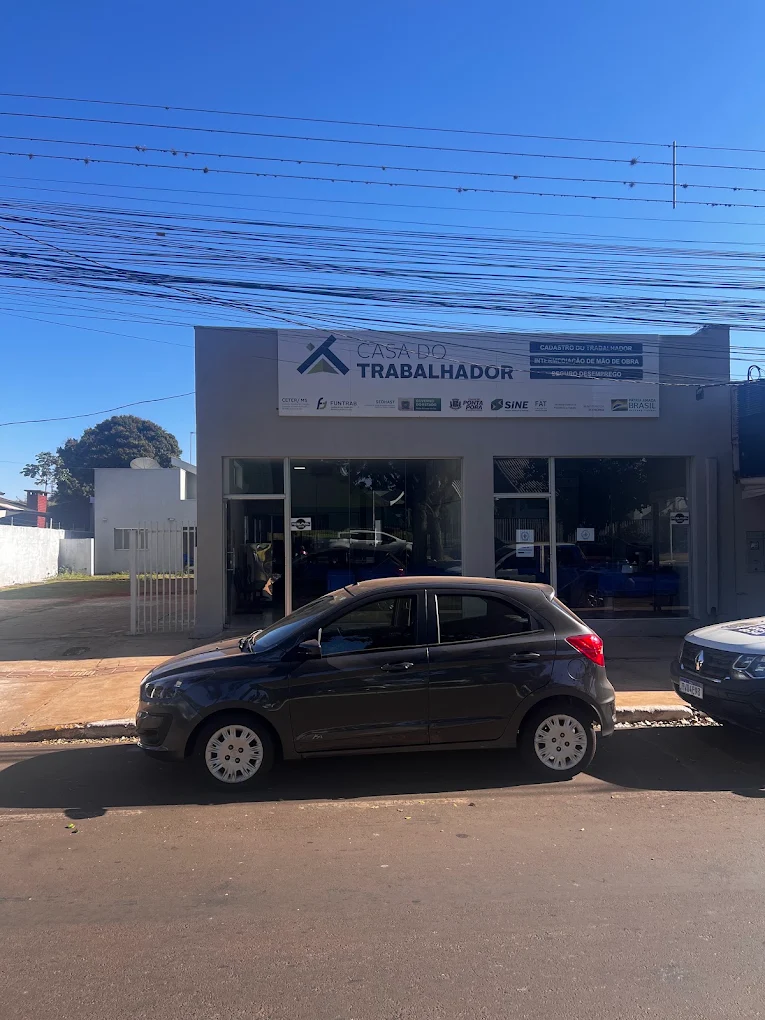 Fachada da Casa do Trabalhador de Ponta Porã, prédio branco de um andar com letreiro na parede e duas frentes de vidro, e um carro escuro estacionado na rua em frente sob céu azul