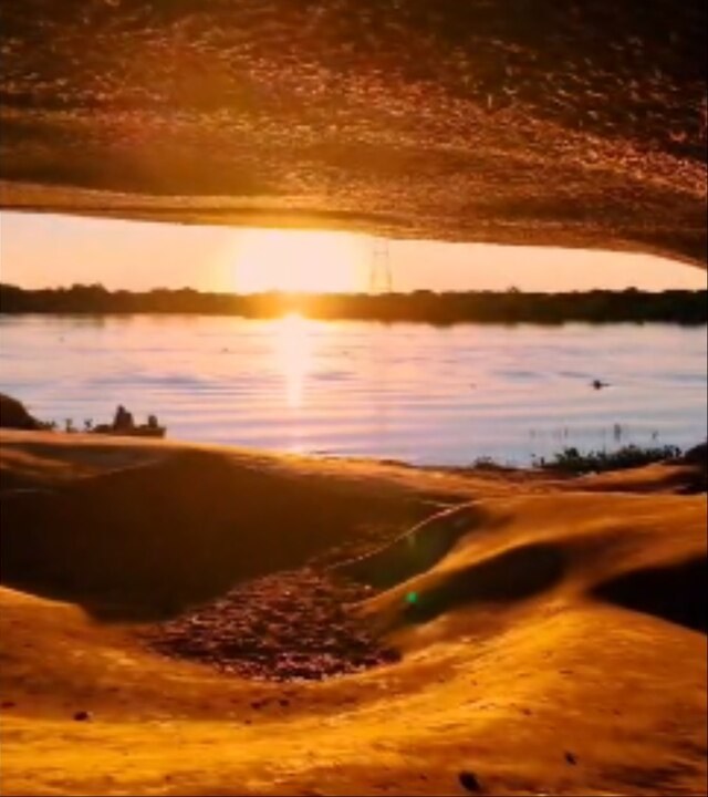 Pôr do sol visto de dentro da caverna: o teto de rocha alaranjada emoldura o sol baixo sobre o Rio Paraguai, com a luz dourada refletida na água e uma faixa de mata ao fundo