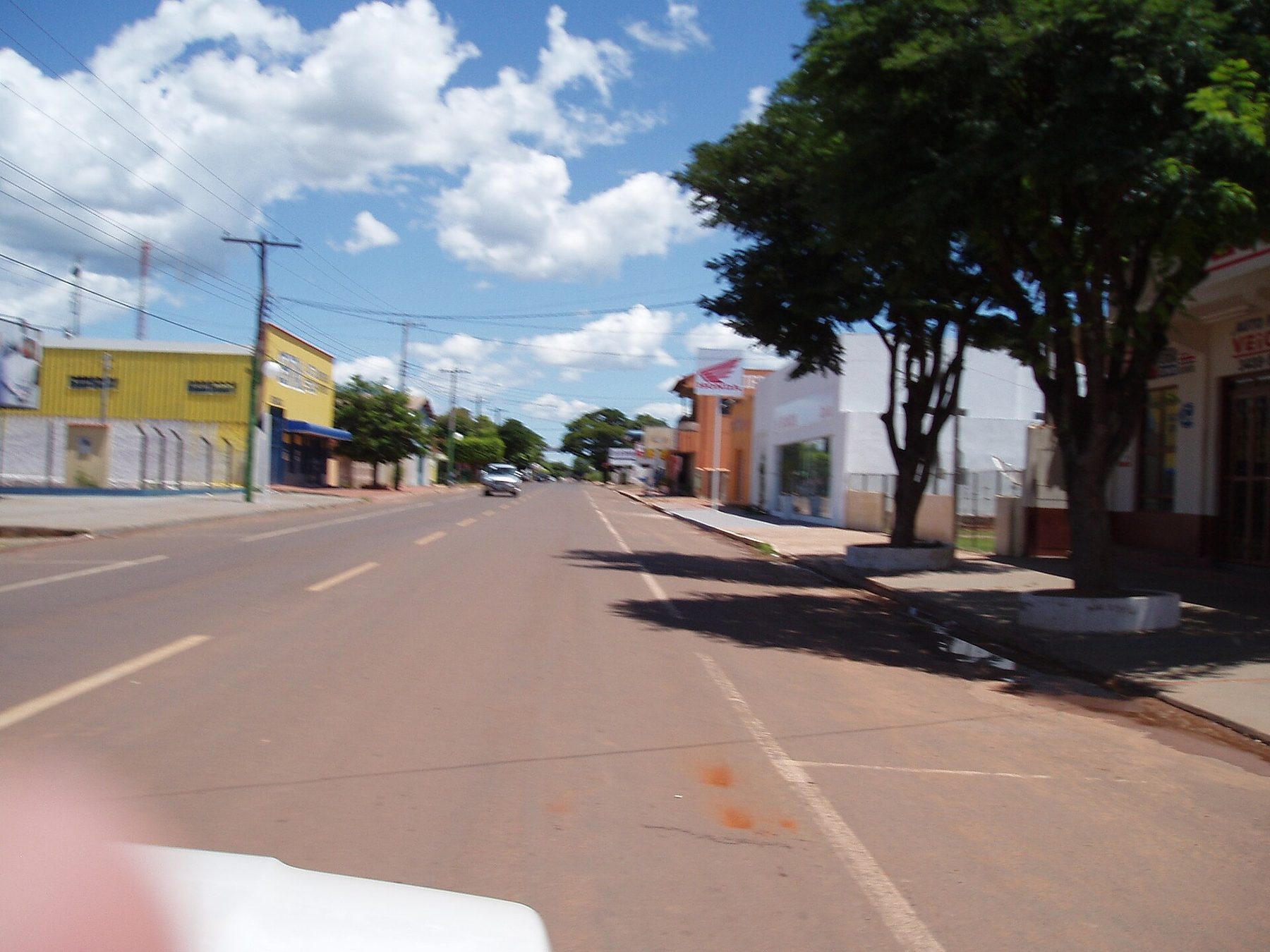Rua comercial de Bela Vista sob céu azul com nuvens, com prédios baixos e uma árvore à direita