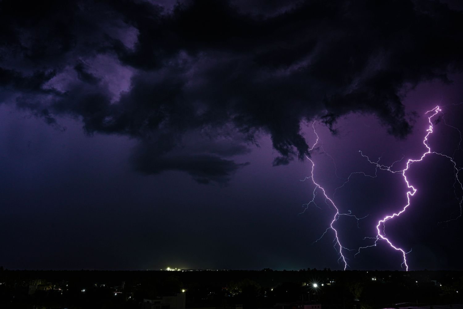 Céu noturno com nuvens escuras e dois raios violeta caindo sobre uma cidade de luzes esparsas no horizonte