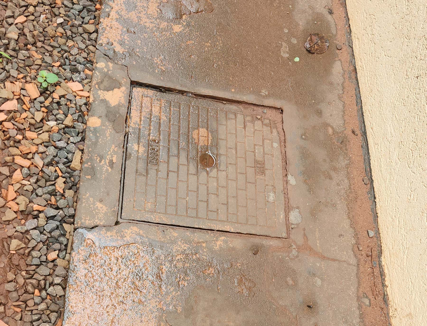 Tampa de caixa de inspeção numa calçada de concreto manchada de terra vermelha, ao lado de pedriscos