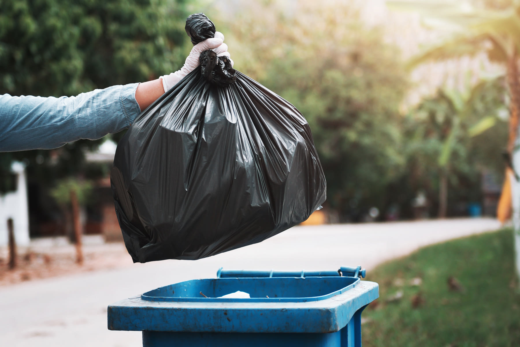 Mano con guante sosteniendo una bolsa de basura negra sobre un contenedor azul abierto, en una calle arbolada.