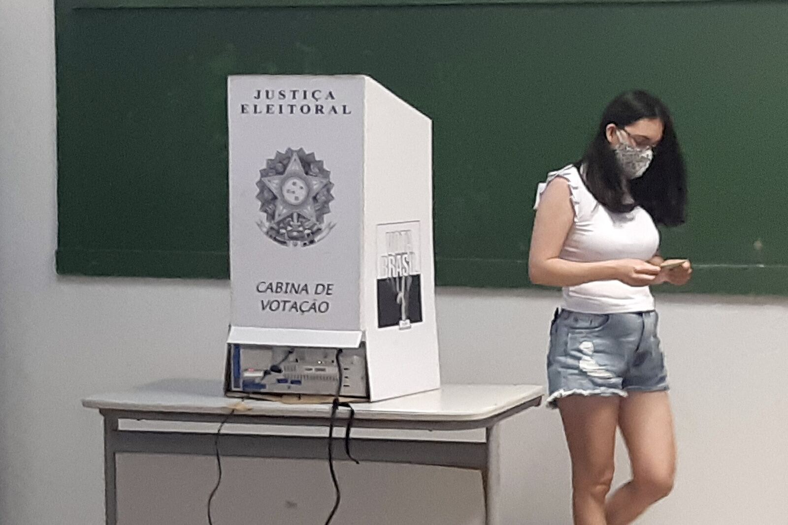 Electora con tapabocas mira el celular al lado de la cabina de votación de la Justicia Electoral brasileña, armada sobre un pupitre escolar