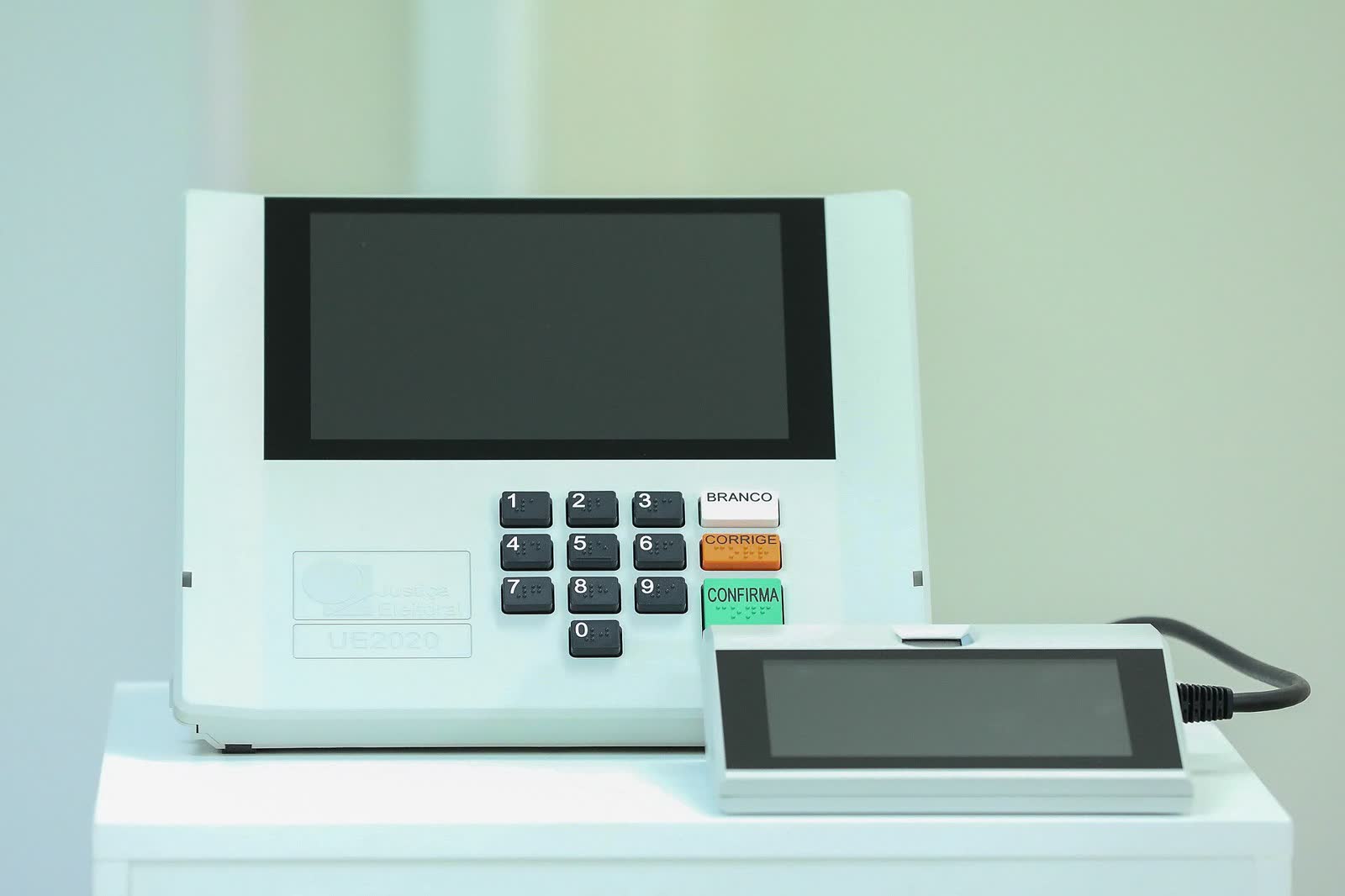 Urna eletrônica brasileira modelo UE2020 sobre uma mesa branca, com teclado numérico e as teclas branco, corrige e confirma à vista, ao lado do terminal do mesário