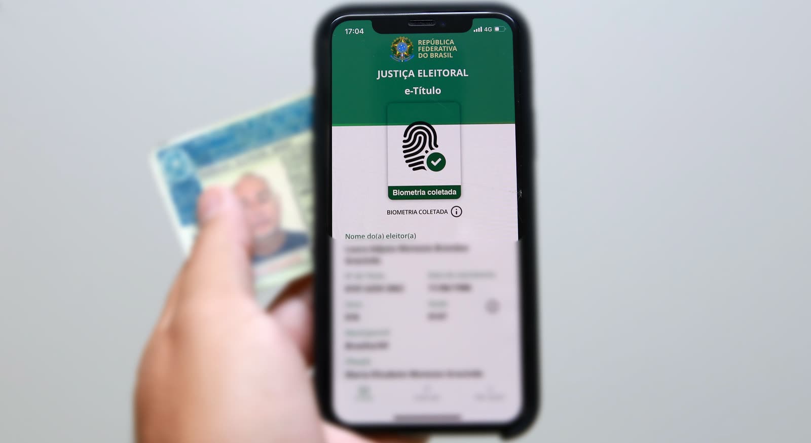 Mão segura um celular com o aplicativo e-Título da Justiça Eleitoral aberto na tela de biometria coletada, com um documento desfocado ao fundo