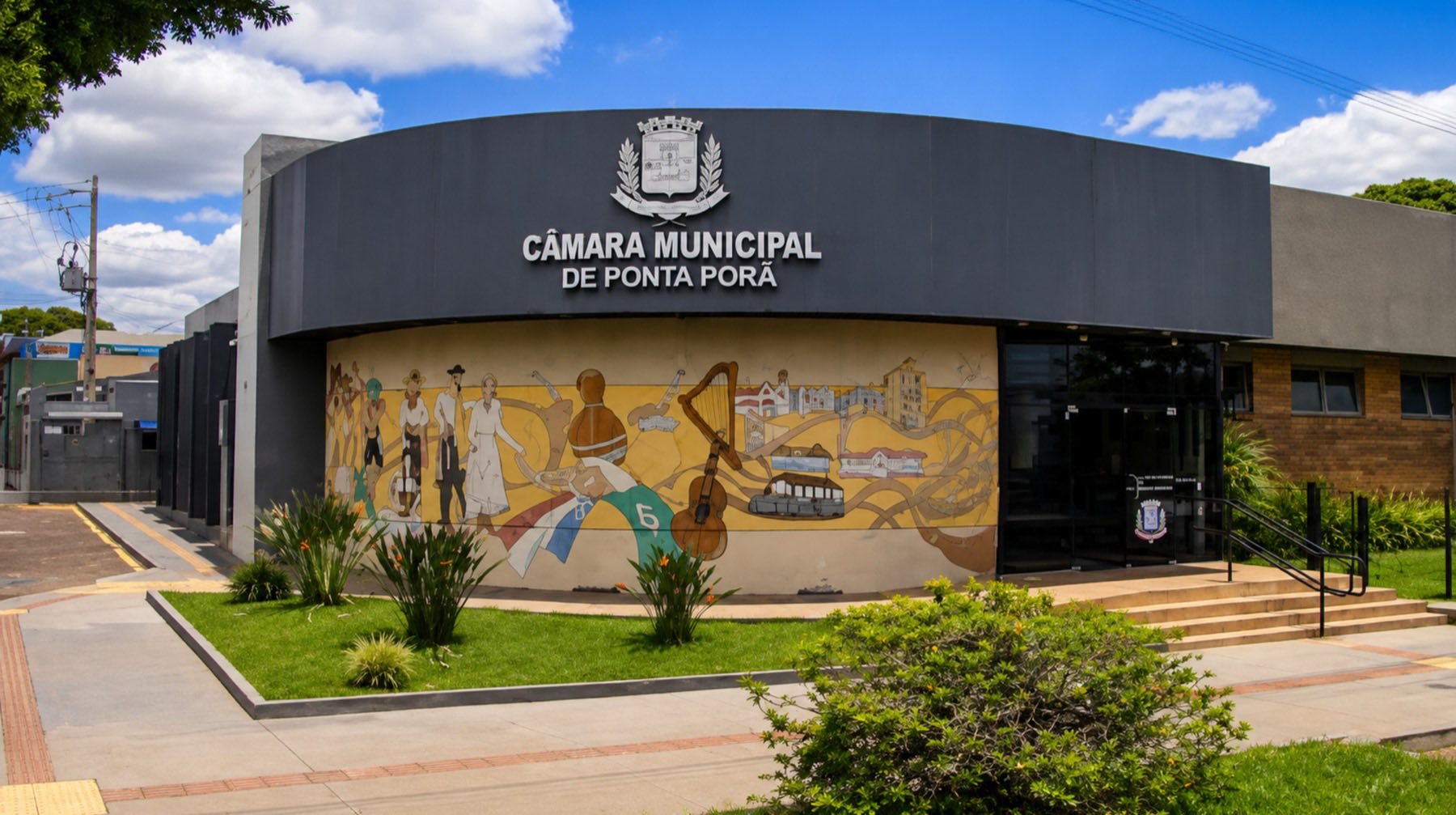 Fachada curva da Câmara Municipal de Ponta Porã, com o brasão do município acima da entrada e um painel colorido na parede