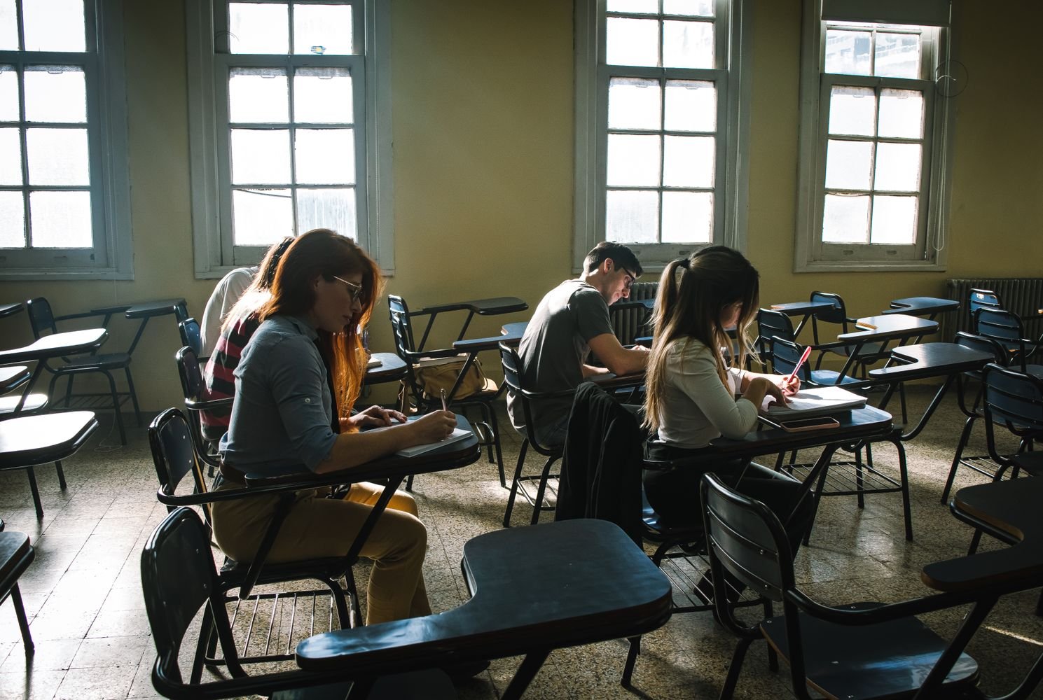 Aula con pupitres escolares, donde cuatro personas sentadas escriben en cuadernos bajo la luz de grandes ventanas