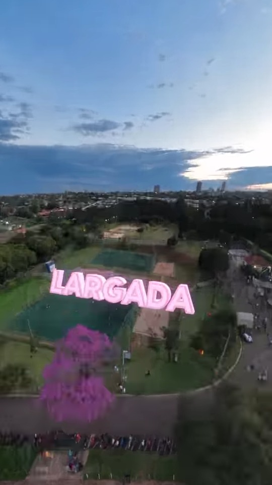 Vista aérea do Horto Florestal ao entardecer, com a palavra 'Largada' em letras lilás sobre a árvore florida junto à pista de largada da corrida
