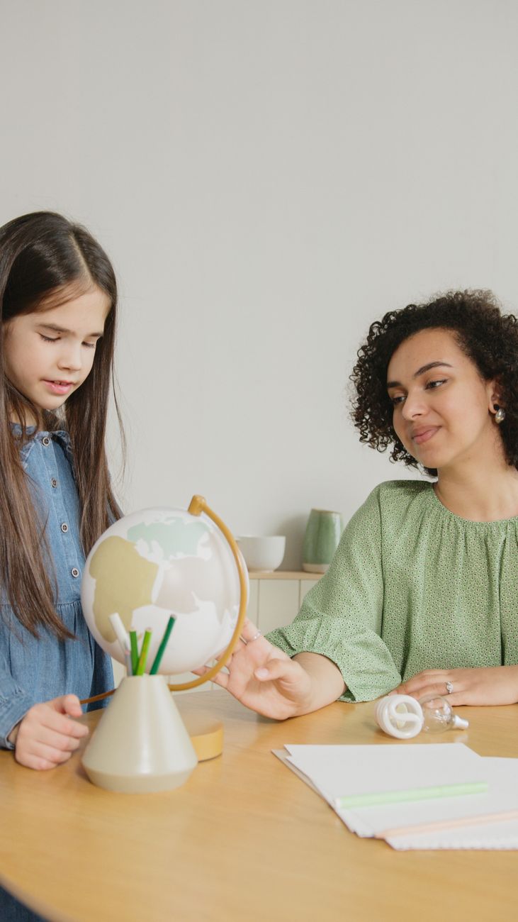 Una niña y una mujer sentadas a una mesa observan juntas un globo terráqueo con lápices de colores
