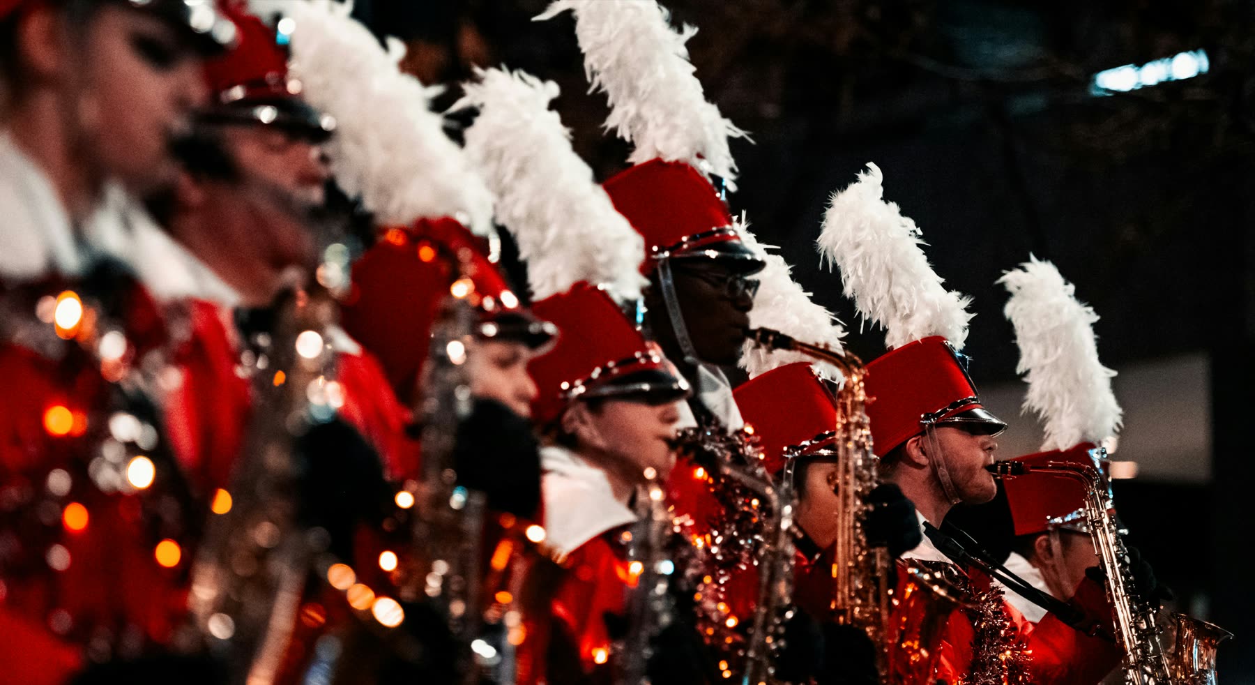 Fila de músicos de banda con uniforme rojo y plumas blancas, tocando saxofón