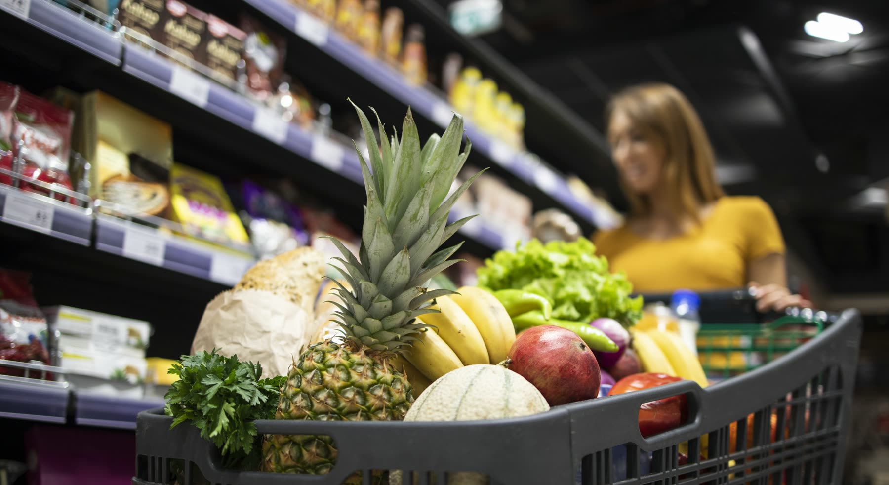 Carrinho de supermercado cheio de frutas, verduras e pão, com prateleiras de produtos ao fundo e uma cliente desfocada empurrando o carrinho.