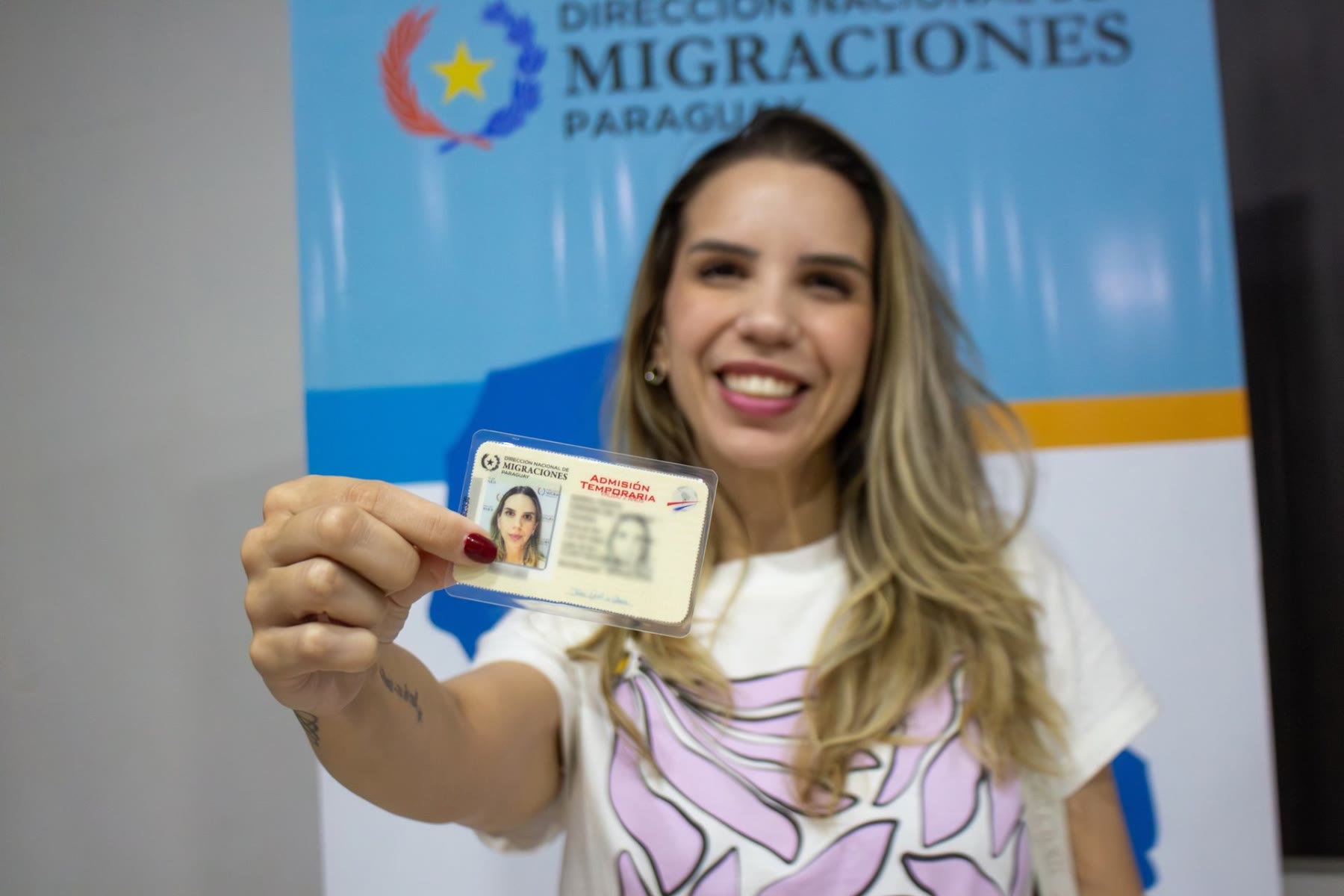 Mujer sostiene frente al rostro una tarjeta de admisión temporaria de la Dirección Nacional de Migraciones del Paraguay, con el banner del organismo al fondo.