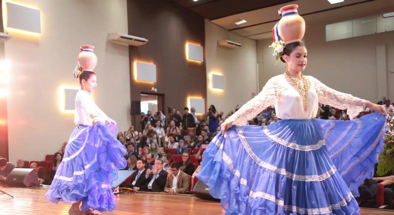 Dos bailarinas giran en el escenario con pollera azul y cántaro en equilibrio sobre la cabeza, frente a una platea llena
