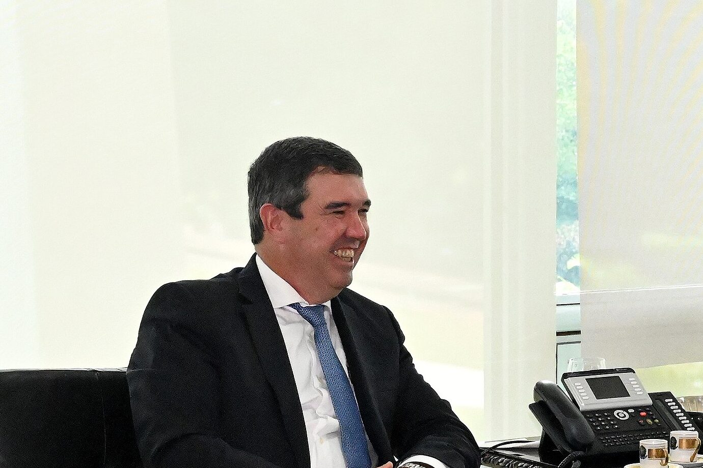 Eduardo Riedel sorri sentado num sofá de couro preto, de terno escuro e gravata azul, numa sala com janelas de vidro