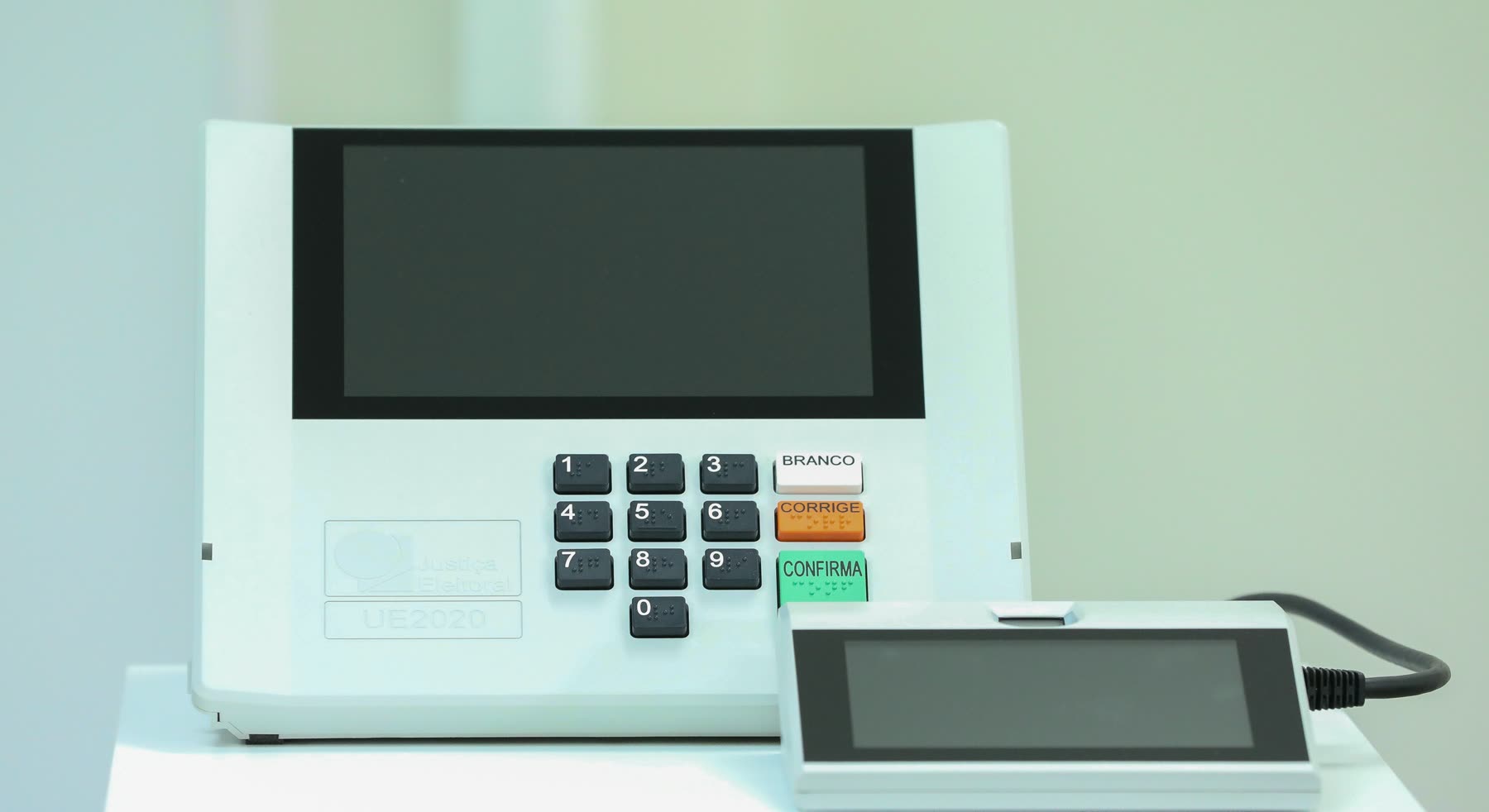 Urna eletrônica brasileira modelo UE2020 com a tela apagada, o teclado numérico e as teclas Branco, Corrige e Confirma, ao lado do terminal do mesário