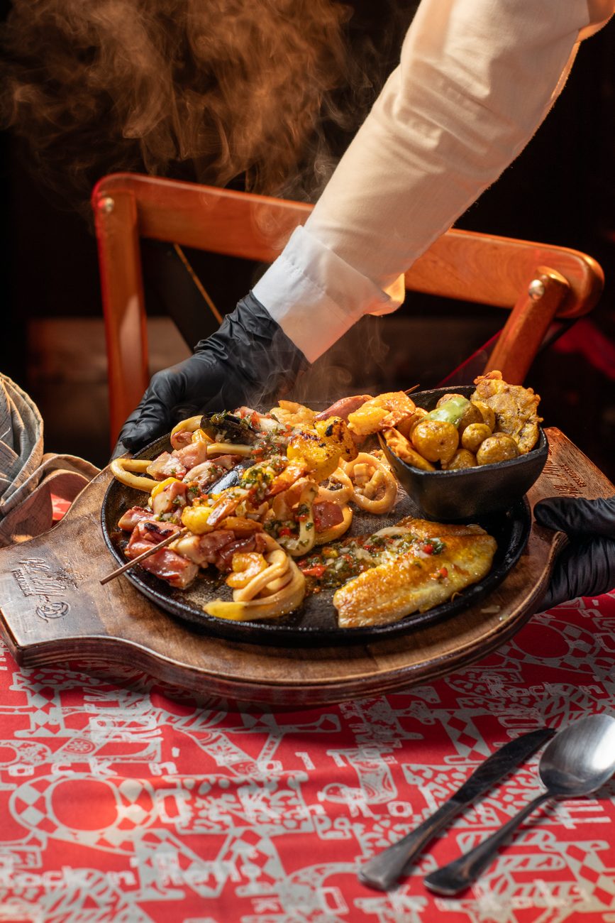 Mão de luva preta serve travessa de ferro com frutos do mar grelhados, peixe e batatas sobre toalha vermelha
