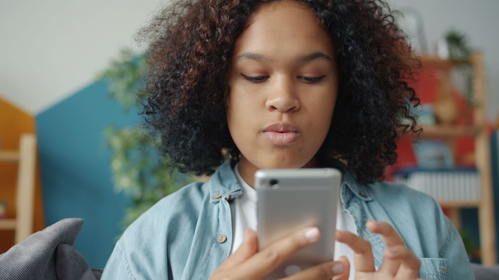 Adolescente de cabelo cacheado e camisa jeans olhando para a tela de um celular que segura com as duas mãos, dentro de casa