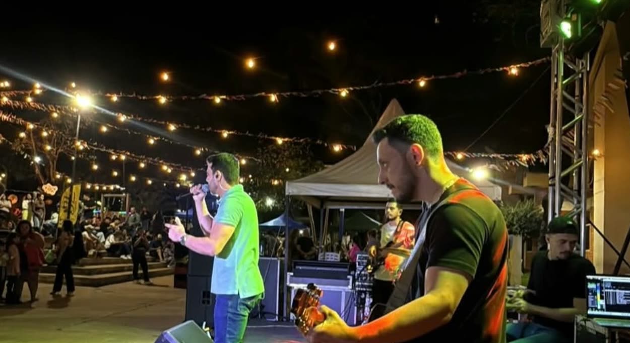 Cantante al micrófono y guitarrista en escenario al aire libre de noche, bajo guirnaldas de luces