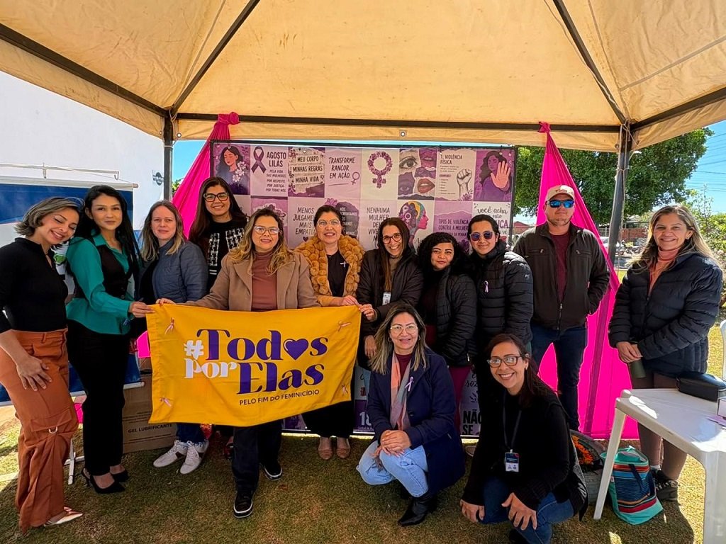 Grupo de mujeres y hombres bajo la Carpa Lila sostiene una bandera amarilla con el lema Todos por Elas, por el fin del feminicidio