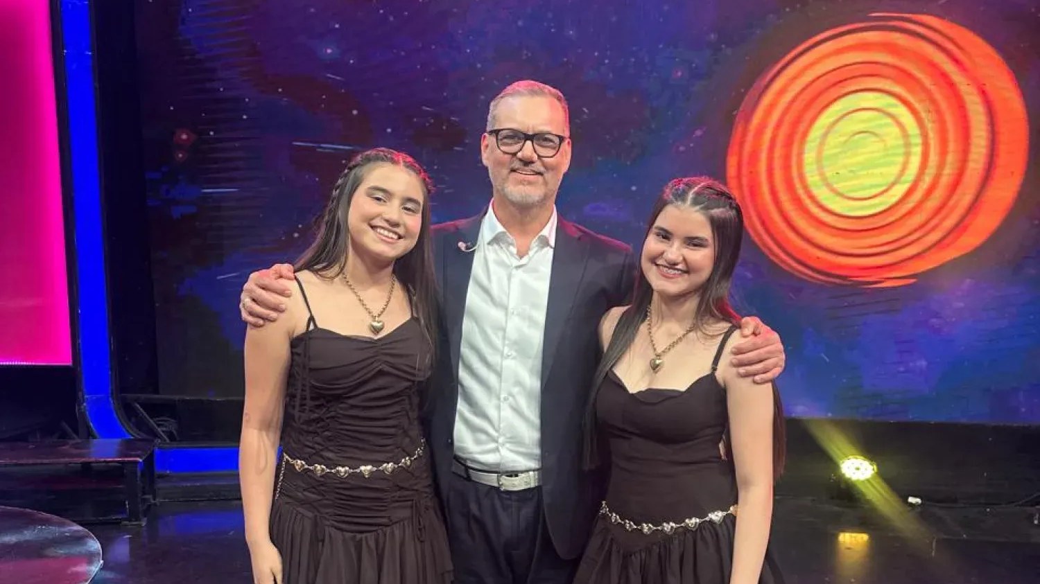 As irmãs Giovanna e Gabrielly Gómez, de vestido preto, posam ao lado do apresentador Palo Rubin no palco do programa El Conejo