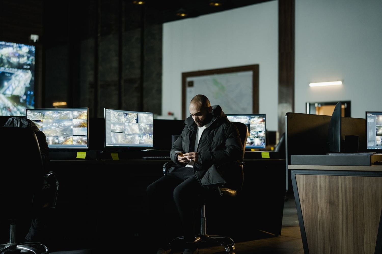 Centro de monitoramento escuro com telas de câmeras de vigilância, um mapa na parede e um homem sentado usando o celular