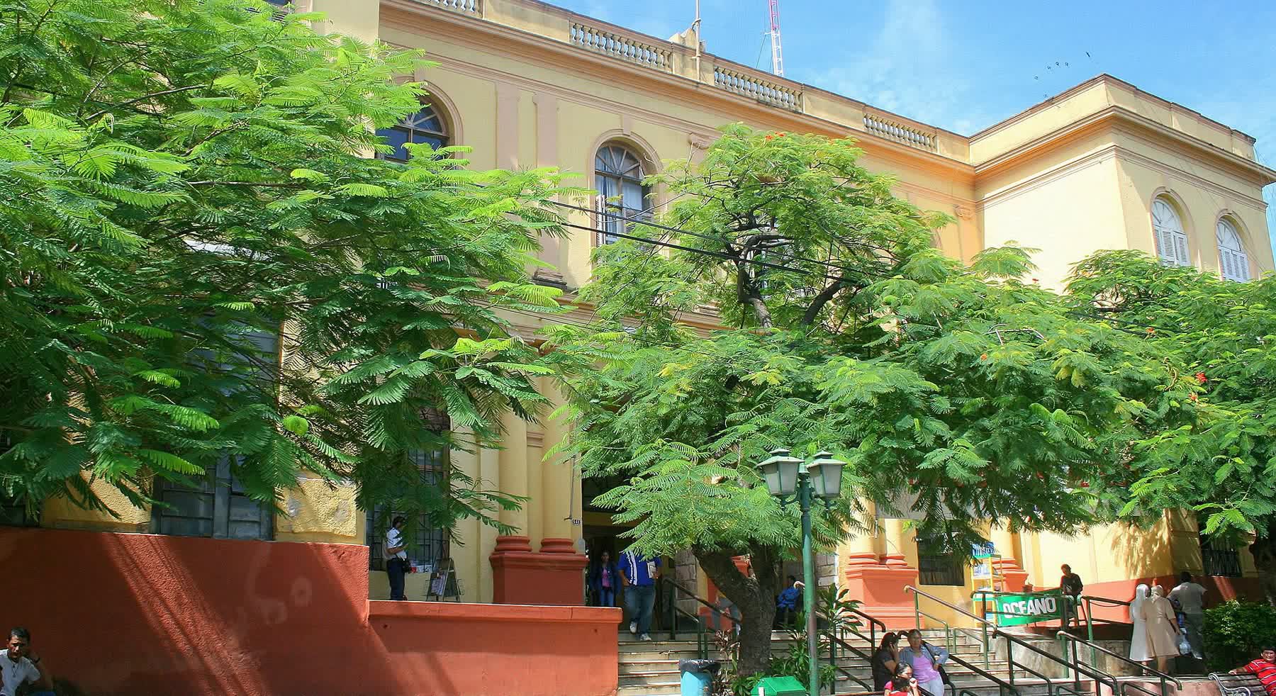 Fachada histórica em tom amarelo do Hospital de Clínicas de Assunção, com árvores na frente e pessoas sentadas na escadaria.