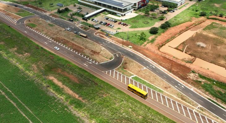 Vista aérea de uma avenida asfaltada com um ônibus escolar amarelo em movimento, com prédios, estacionamento e área verde ao lado da pista.