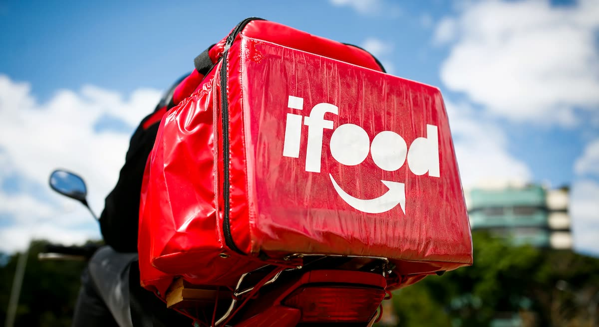 Bolsa térmica vermelha do iFood na garupa de uma moto de entrega, com céu azul e nuvens ao fundo