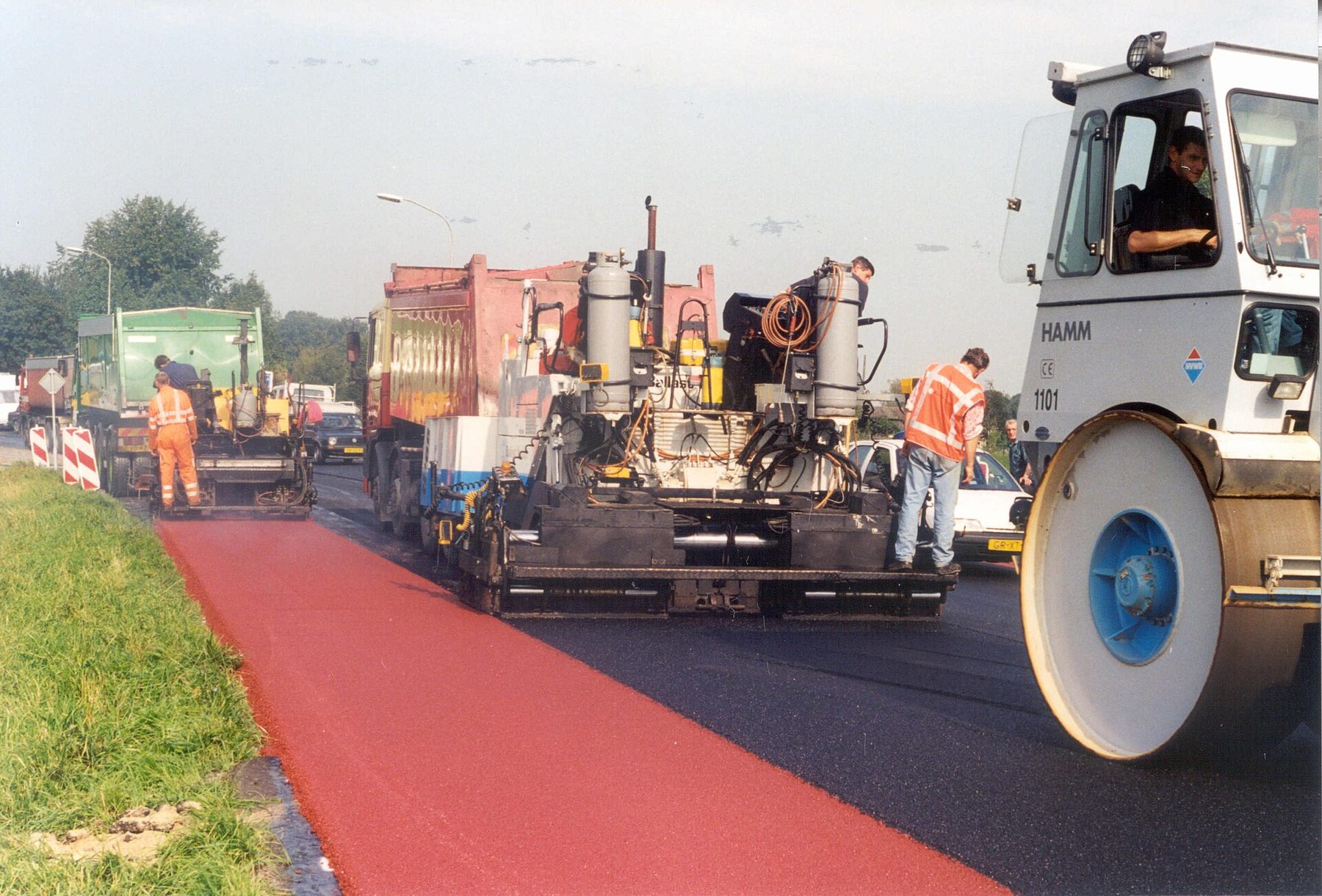 Máquinas e operários aplicam asfalto novo numa rua, com uma faixa vermelha ao lado do gramado e um rolo compactador em primeiro plano