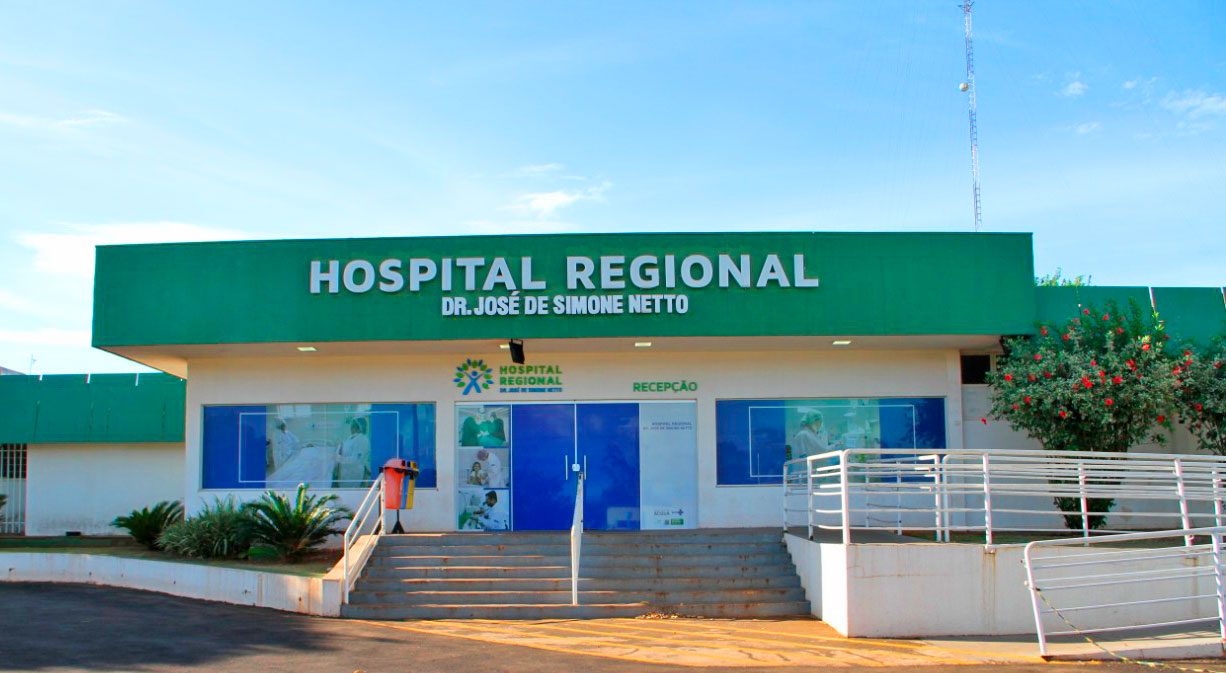 Fachada verde e branca do Hospital Regional de Ponta Porã, com a escada e a rampa de acesso à recepção e um arbusto florido à direita