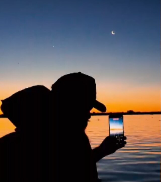 Silhuetas de duas pessoas de boné contra o céu do entardecer, uma delas fotografando o pôr do sol com o celular, com a lua crescente no alto e o rio ao fundo