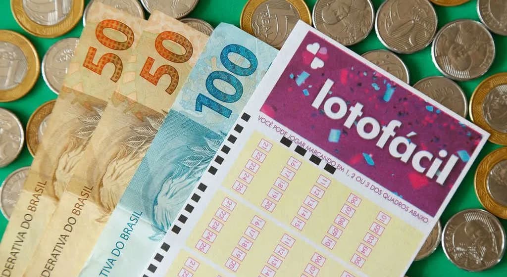 Volante da Lotofácil por cima de notas de R$ 50 e de R$ 100 e de moedas de real, sobre fundo verde