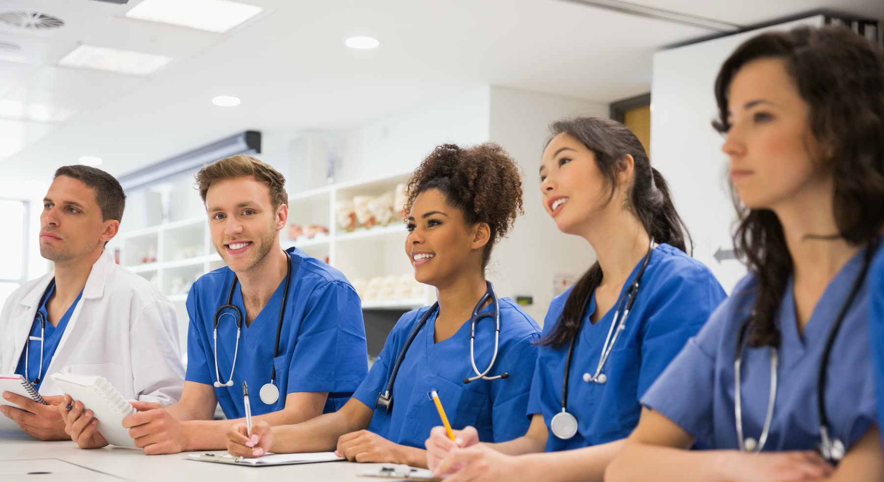 Cinco estudantes de medicina com pijama cirúrgico azul e estetoscópio no pescoço, sentados lado a lado numa bancada de sala de aula, anotando.