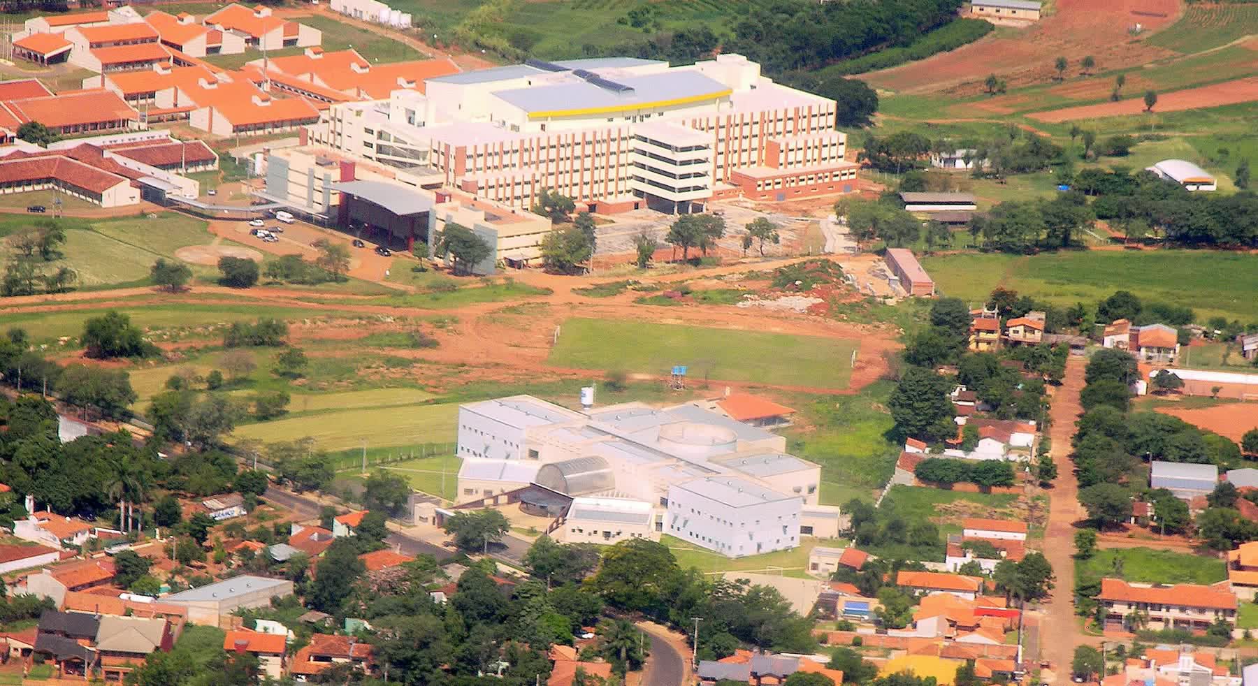 Vista aérea de um complexo hospitalar universitário, com prédios claros de vários andares cercados por área verde e estacionamento.