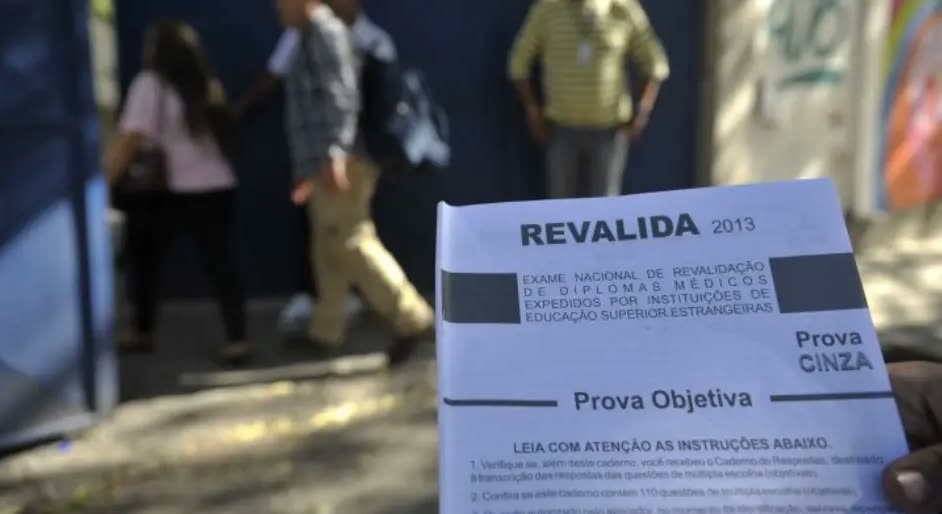Caderno de prova do Revalida segurado à frente da câmera, com candidatos desfocados entrando no local de prova ao fundo.