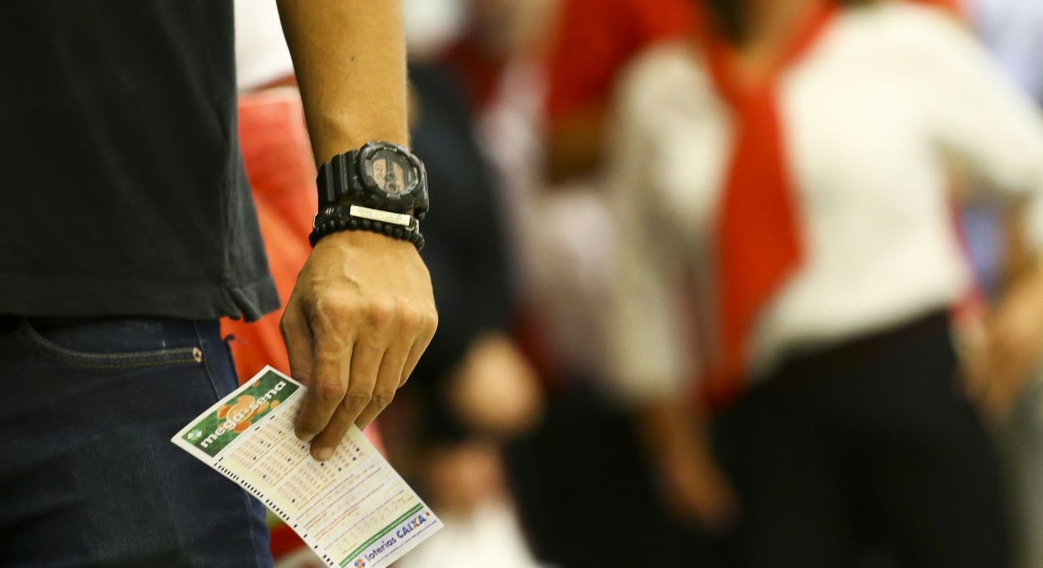 Mano de un apostador sosteniendo un volante de la Mega-Sena, con personas en fila desenfocadas al fondo en una casa de lotería