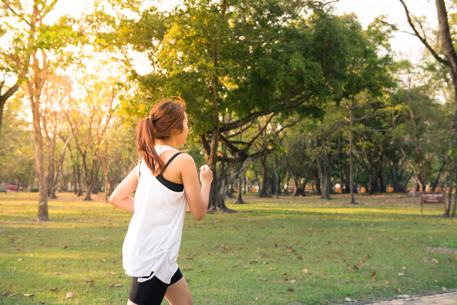 Mujer con blusa blanca sin mangas corre en un parque arbolado, con la luz baja del sol entre los árboles