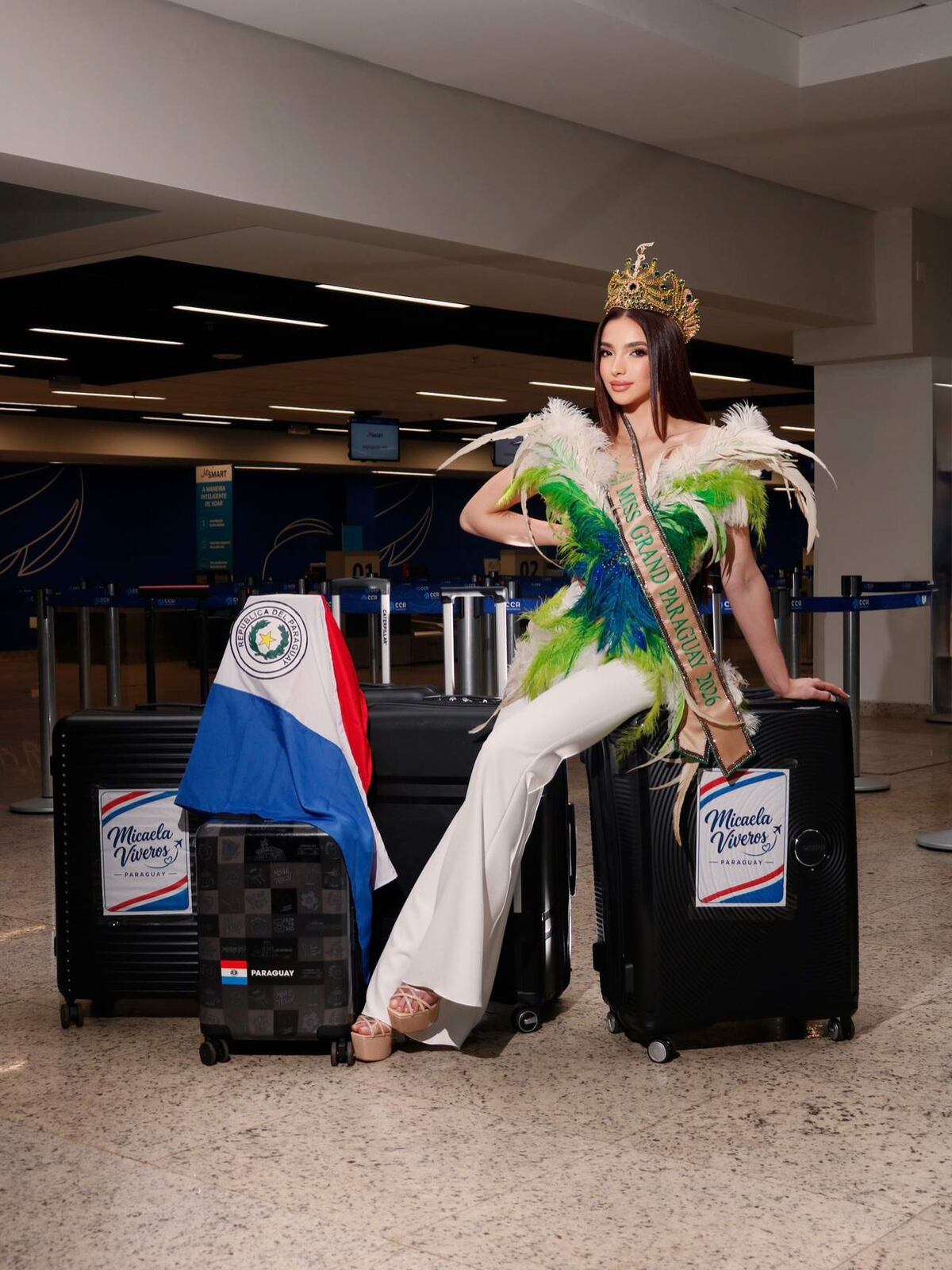 Micaela Viveros con corona y banda de Miss Grand Paraguay 2026, blusa de plumas verdes, azules y blancas, sentada sobre una valija negra en el hall de un aeropuerto, junto a otras valijas y la bandera de Paraguay