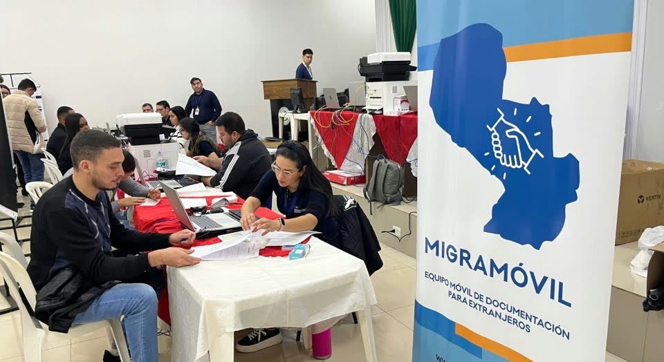 Atención en mesas con documentos y computadoras, junto al banner del Migramóvil