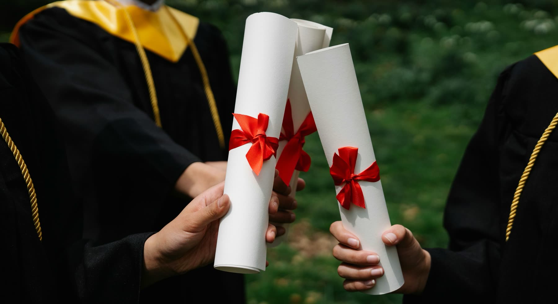Manos de recién graduados sostienen diplomas enrollados con cinta roja en una ceremonia de graduación al aire libre