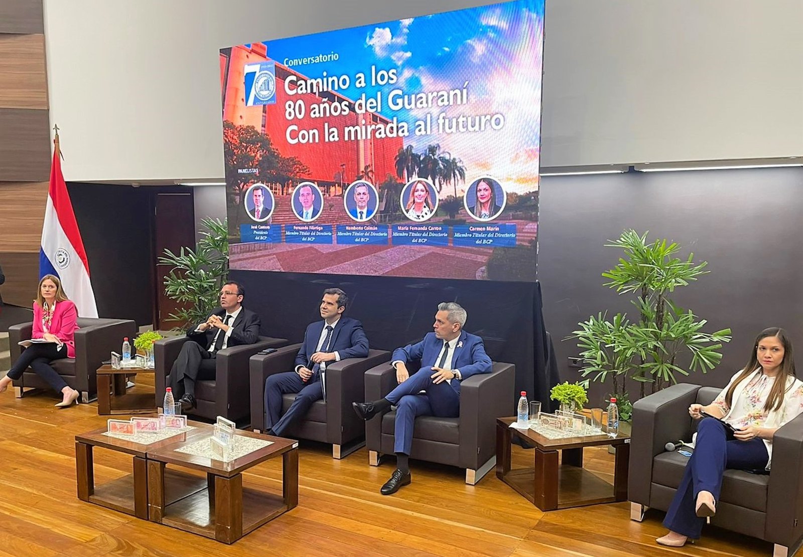 Diretores do Banco Central do Paraguai sentados em poltronas no palco de um conversatório, com um painel escrito Camino a los 80 años del Guaraní ao fundo e cédulas expostas nas mesinhas à frente